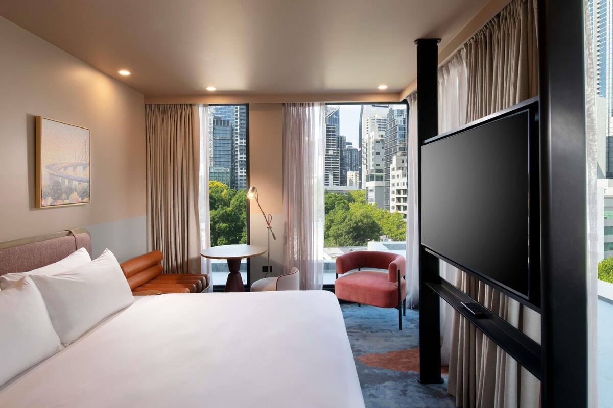A hotel room with a large bed, a TV, a desk, a chair, and a view of the cityscape.