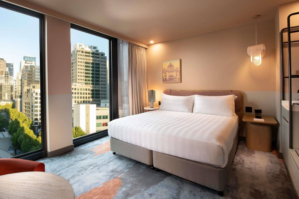 A hotel room with a large bed, a nightstand, a lamp, a view of the cityscape, and a window with curtains.