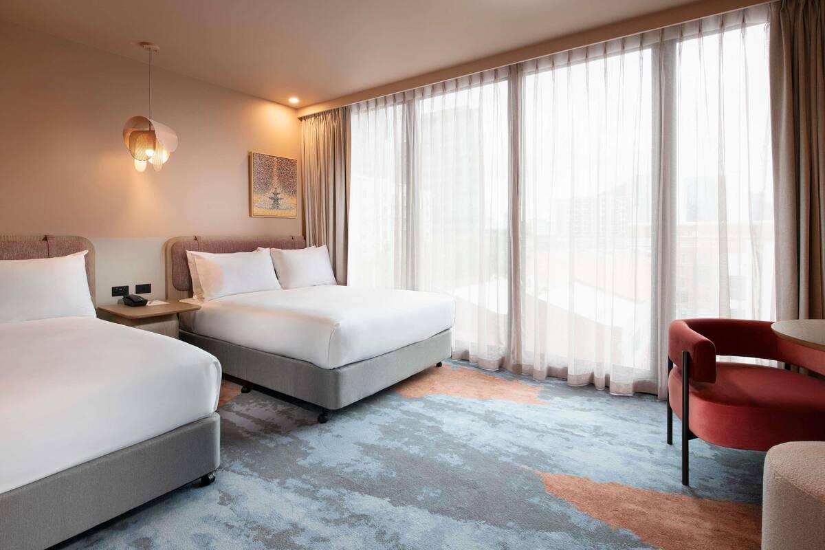A hotel room with two beds, a desk, a chair, and a large window with curtains.