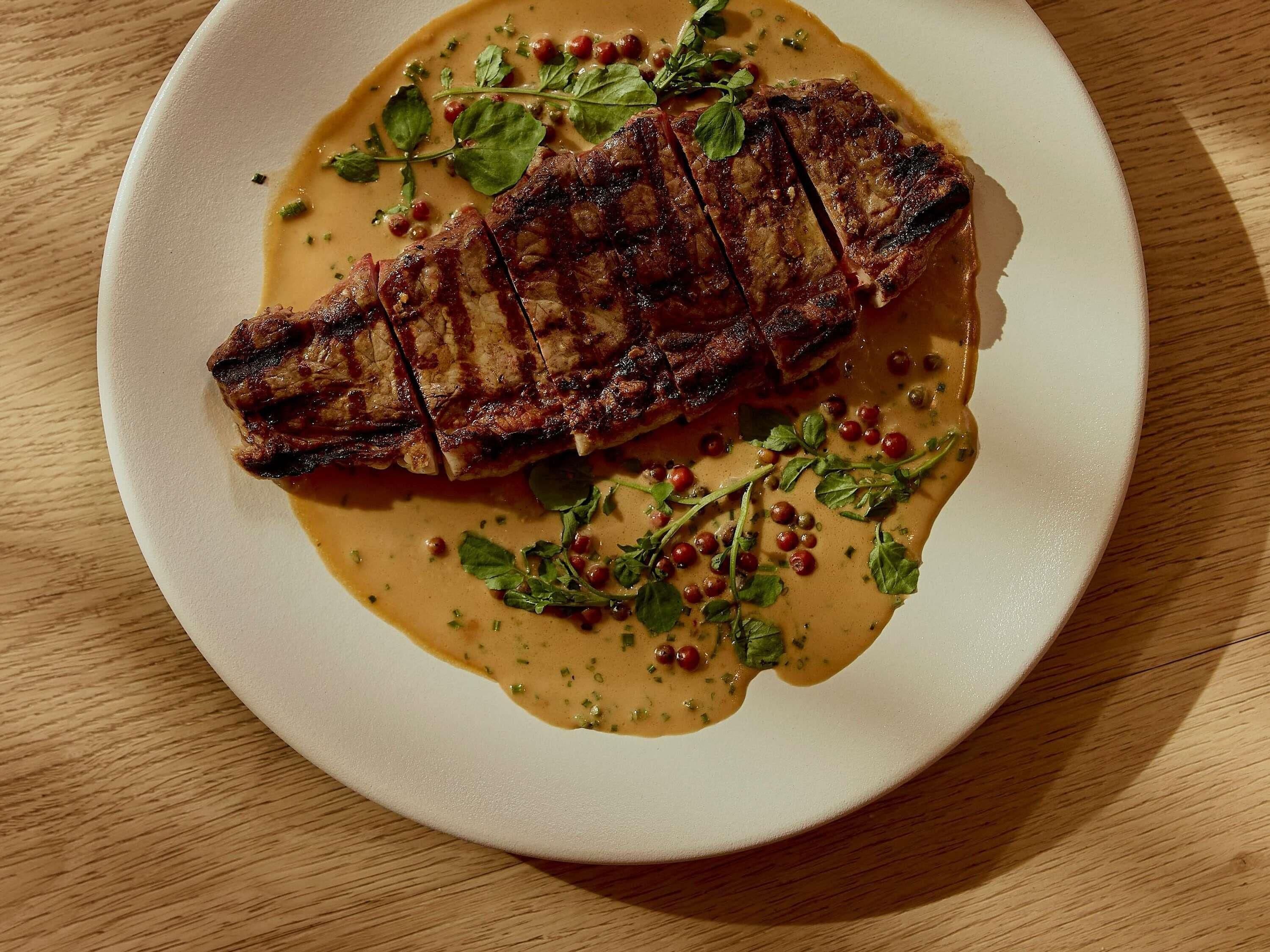 Une assiette blanche sur laquelle est servi un steak grillé, agrémenté d’herbes et de grains de poivre rouge.