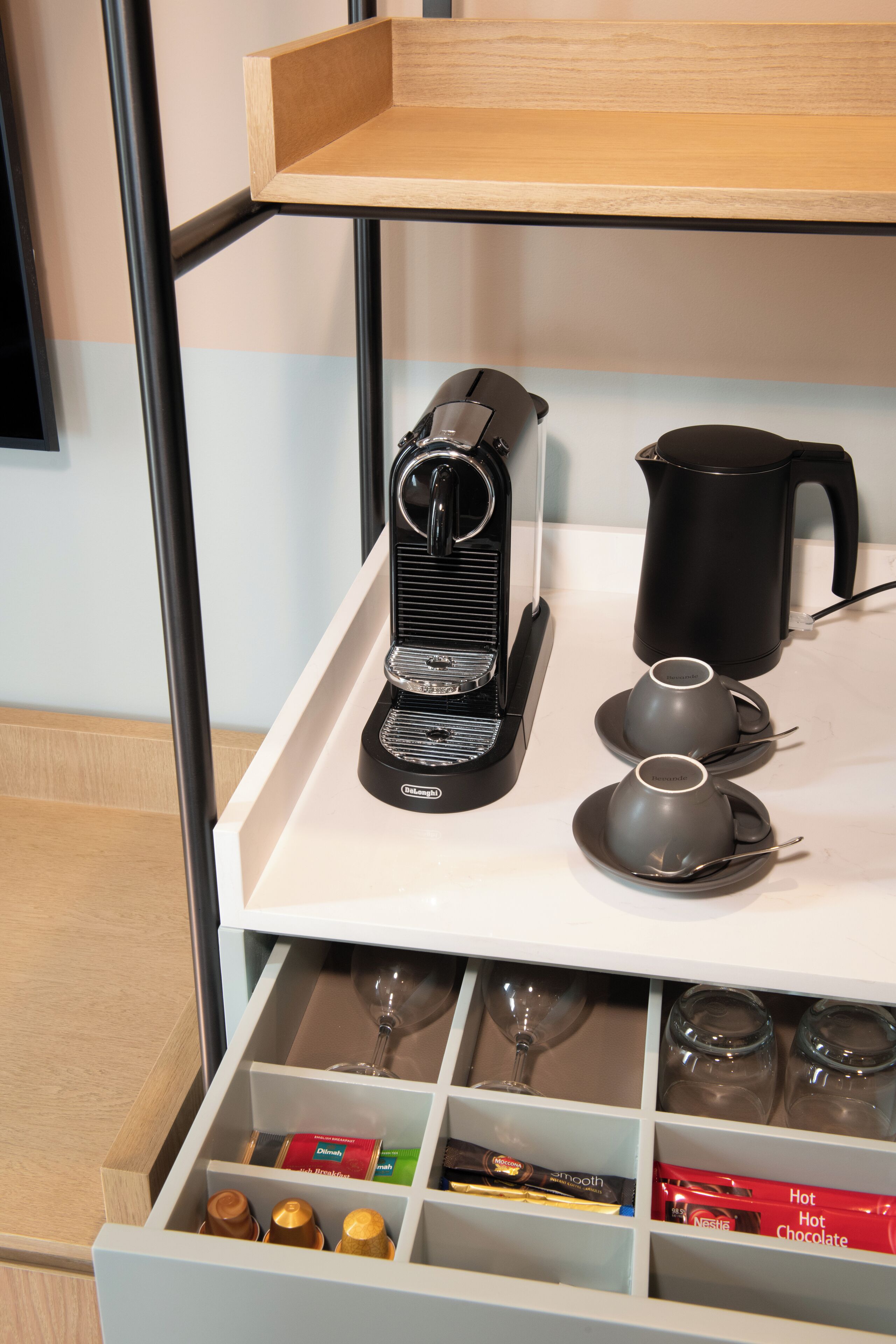 Una cafetera, un hervidor y dos tazas sobre una bandeja. Abajo, hay compartimentos con vasos de vino, barras de chocolate y una caja con la etiqueta 'Chocolate Caliente'.