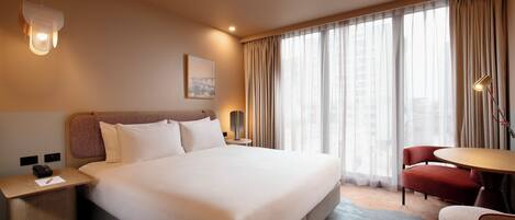 A hotel room with a large bed, a desk with a lamp, a chair, and a view of the city through the window.