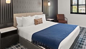 Ein modernes Hotelzimmer mit einem großen Bett, einem Nachttisch, einem Sessel und einem Fenster mit Jalousien.