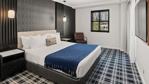 Ein modernes Hotelzimmer mit einem großen Bett, einem Nachttisch, einem Sessel, einem Fenster mit Jalousien und einem Fernseher.