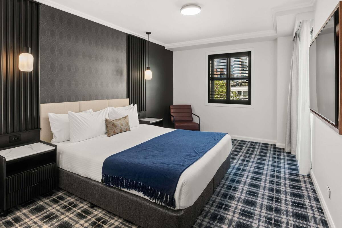 Habitación de hotel moderna con una cama grande, una mesita de noche, una silla, una ventana con persianas y un televisor.