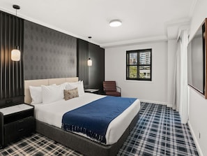 Une chambre d’hôtel moderne avec un grand lit, une table de chevet, une chaise, une fenêtre avec des stores et un téléviseur.