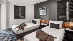 Ein Hotelzimmer mit zwei Betten, einem großen Fenster mit Jalousien, einem gerahmten Kunstwerk an der Wand und einem Nachttisch mit Lampe.