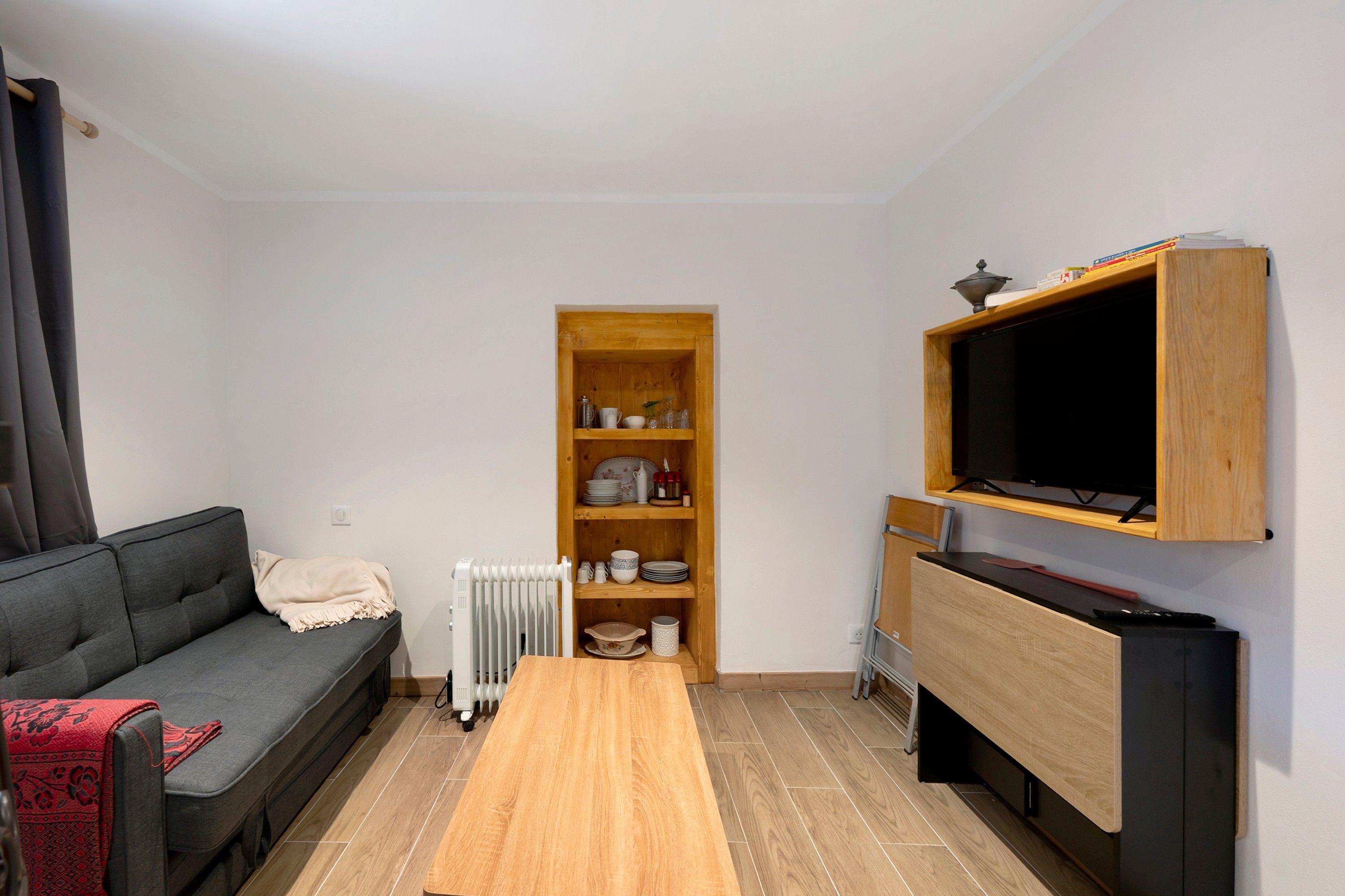 Een woonkamer met een grijze bank, een houten salontafel, een aan een houten voetstuk bevestigde televisie, en een boekenkast met diverse spullen.