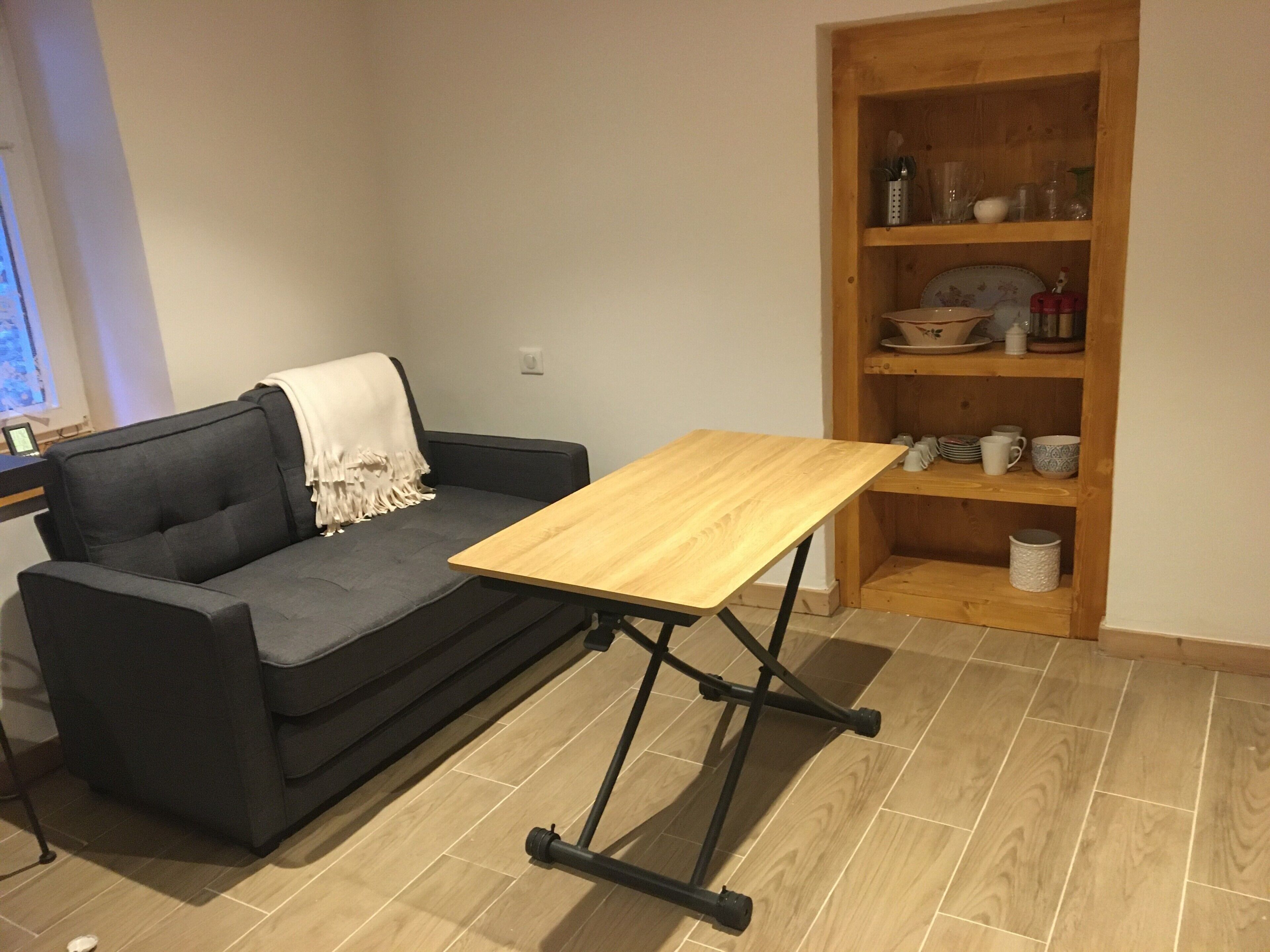 Een kamer met een bank, een klein houten tafeltje, een plank met servies en een betegelde vloer.