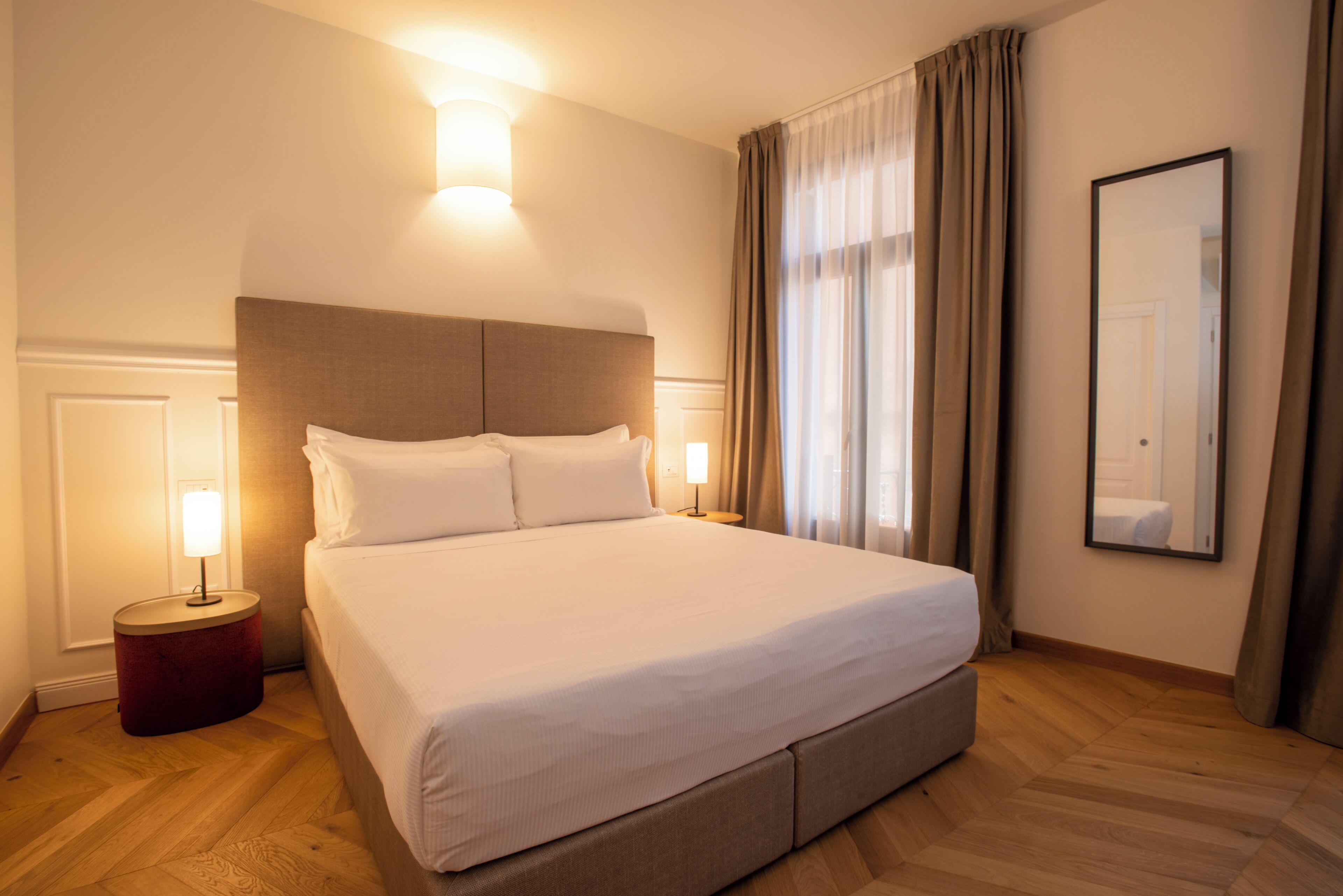Quarto de hotel com uma cama grande, uma mesa de apoio redonda, uma mesinha de cabeceira, um espelho fixado na parede e uma janela com cortinas.