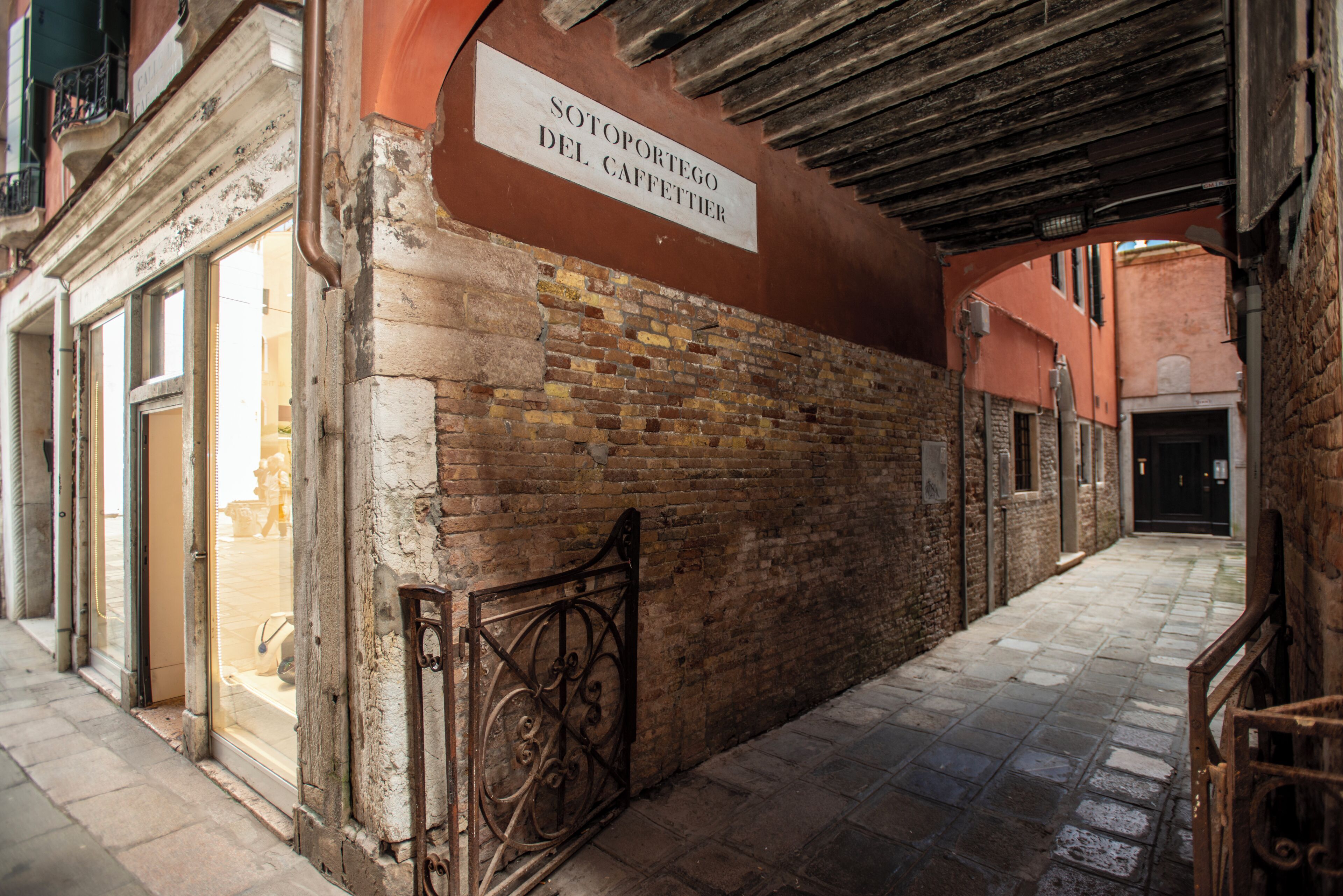 Une ruelle étroite, avec une enseigne indiquant « Sotoportego del Caffetterio », des murs en briques et un portail en métal.