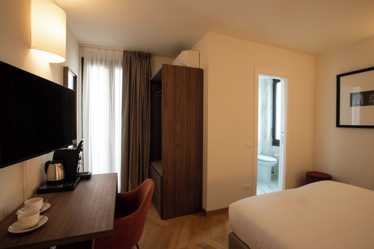 A hotel room with a bed, a desk with a coffee maker, a TV, and a bathroom visible through an open door.