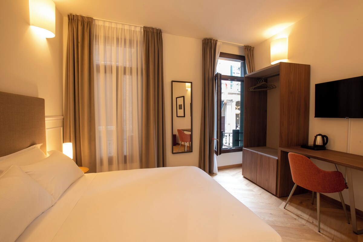 A hotel room with a bed, a desk, a chair, a television, and a view of the outside.
