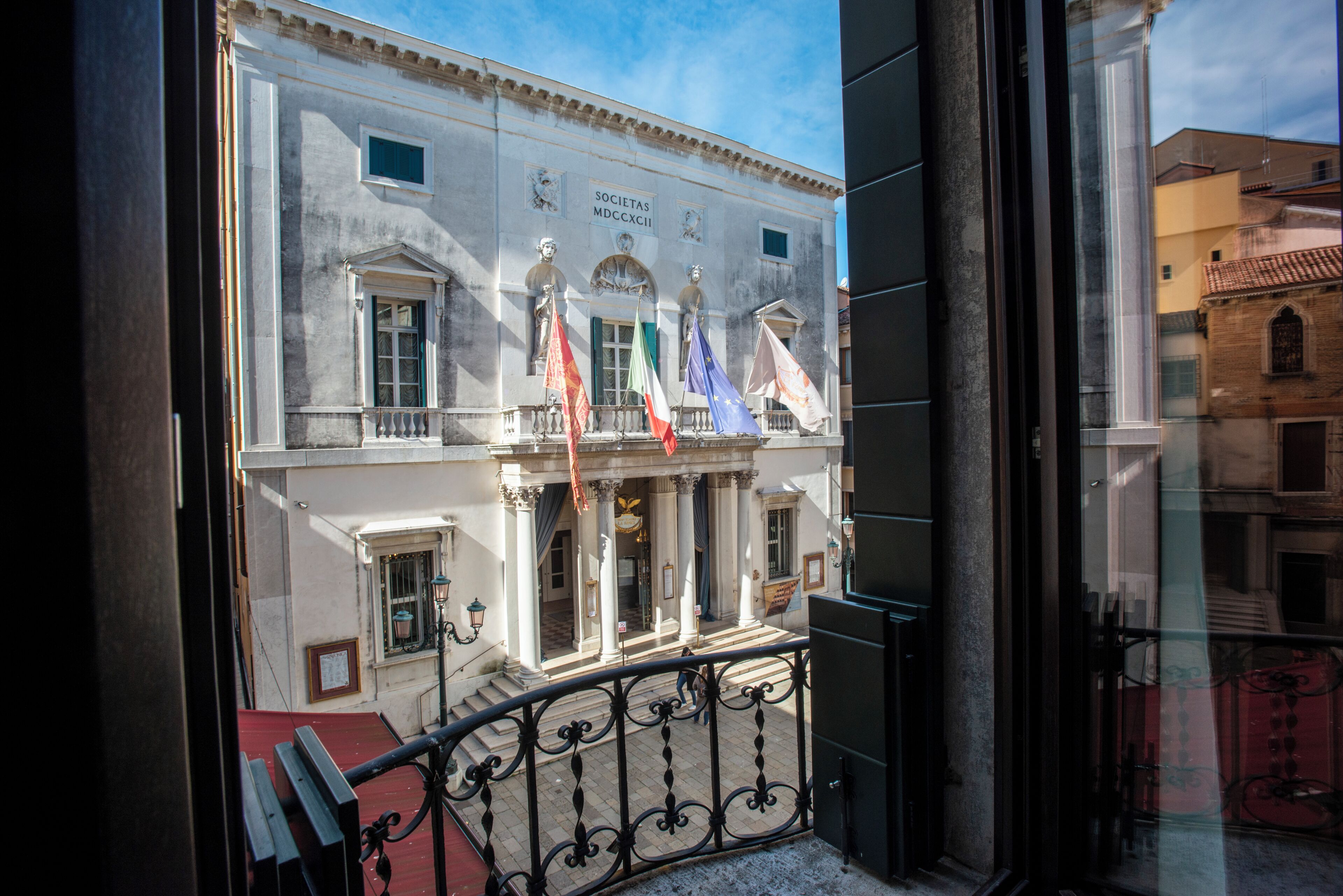 A building with classical architecture, featuring columns, a balcony, and flags.