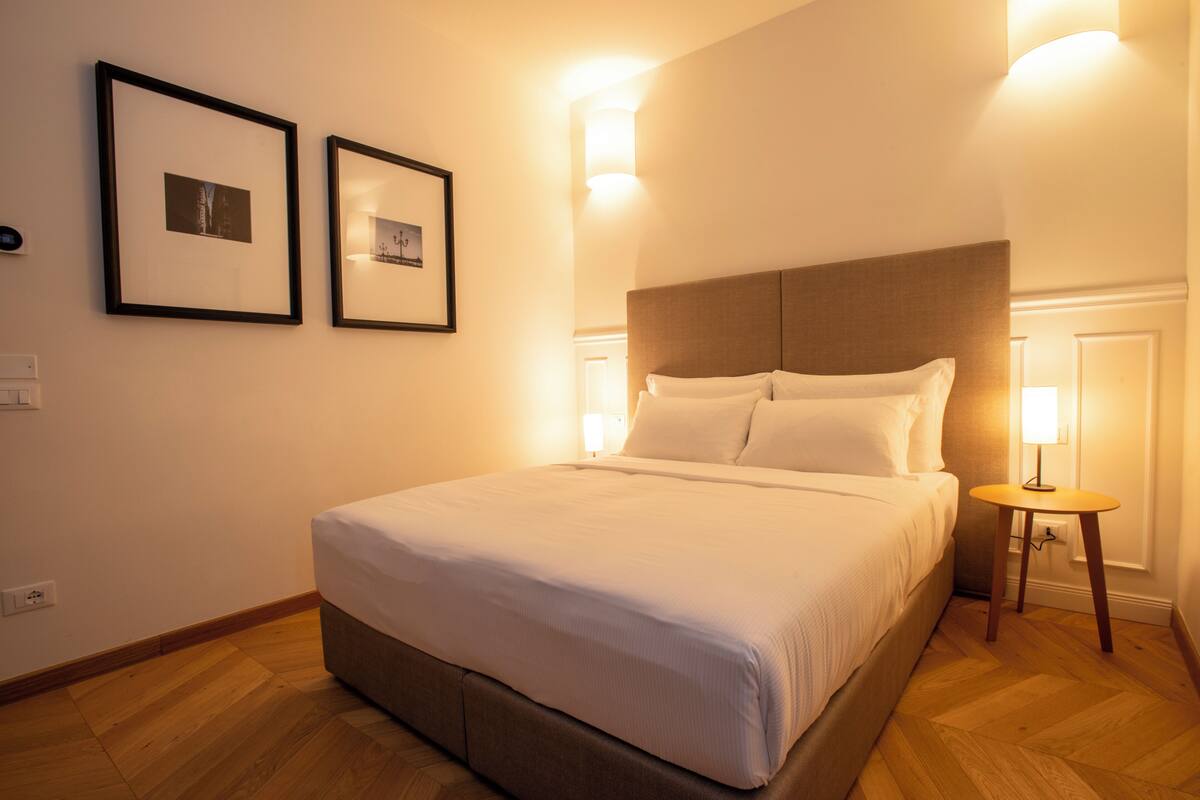 A neatly made bed in a room with two framed pictures on the wall, a bedside table with a lamp, and a wooden floor.