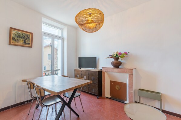 Ein Essbereich mit Tisch und Stühlen, einem Fernseher auf einer Kommode, einem Kamin mit Blumenvase und einem Fenster mit Blick auf Gebäude.