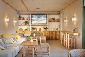 Un espace de vie chaleureux et bien éclairé, avec un banc en bois, un canapé agrémenté de coussins et une table basse. On y trouve des étagères avec des plantes et des objets décoratifs, ainsi qu’une grande fenêtre donnant sur l’extérieur.