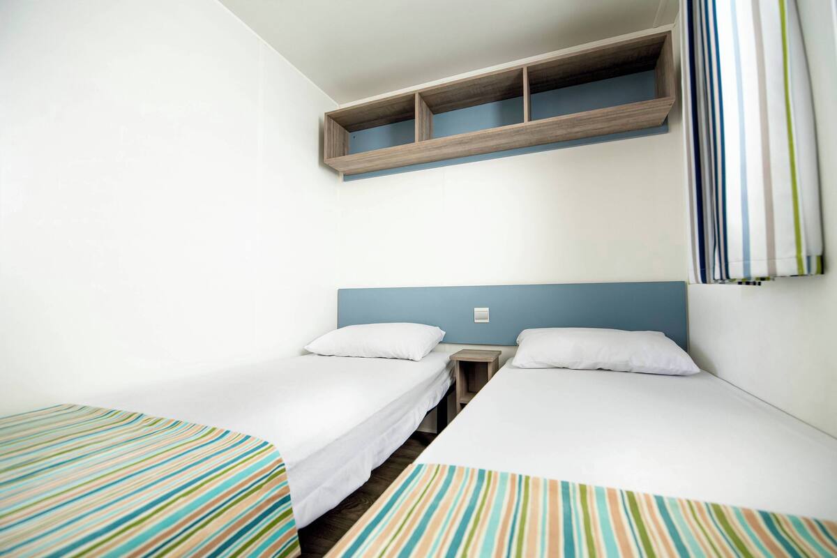 Een kamer met twee eenpersoonsbedden, een houten plank erboven en een blauw hoofdeinde.