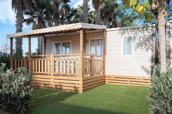 A small, single-story wooden structure with a porch and a covered seating area, surrounded by a well-maintained lawn and palm trees. - Creixell Camping & Family Resort (Creixell)