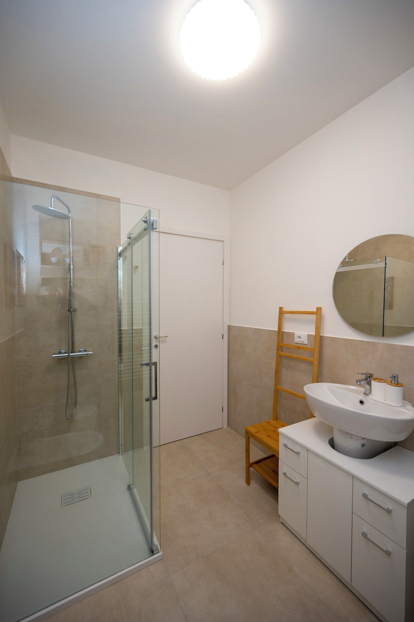 A modern bathroom with a glass shower enclosure, a wooden chair, a sink with a mirror, and a white cabinet.