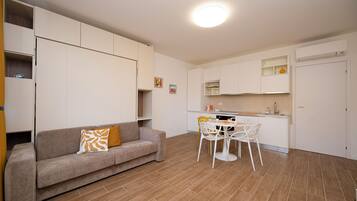 Ein kompakter Wohnraum mit einer Couch, einem Esstisch und Stühlen. Es gibt eine Küchenzeile mit Schränken und einem kleinen Kühlschrank.
