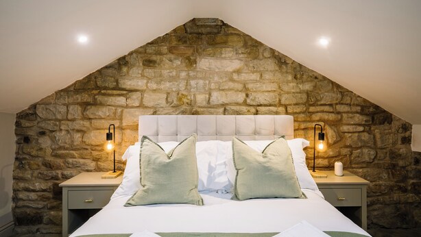 A bedroom with a stone wall, a bed with white bedding, two bedside tables with lamps, and a green throw blanket on the bed. - Sun St Apartment 5 (Lancaster)
