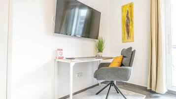 Una habitación moderna con televisión de pantalla plana en la pared, un escritorio con una silla, una pequeña planta en maceta y un cuadro.