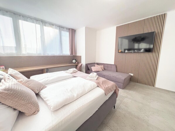 Une chambre à coucher moderne, avec un lit, un canapé, un téléviseur fixé au mur et une fenêtre avec des rideaux.