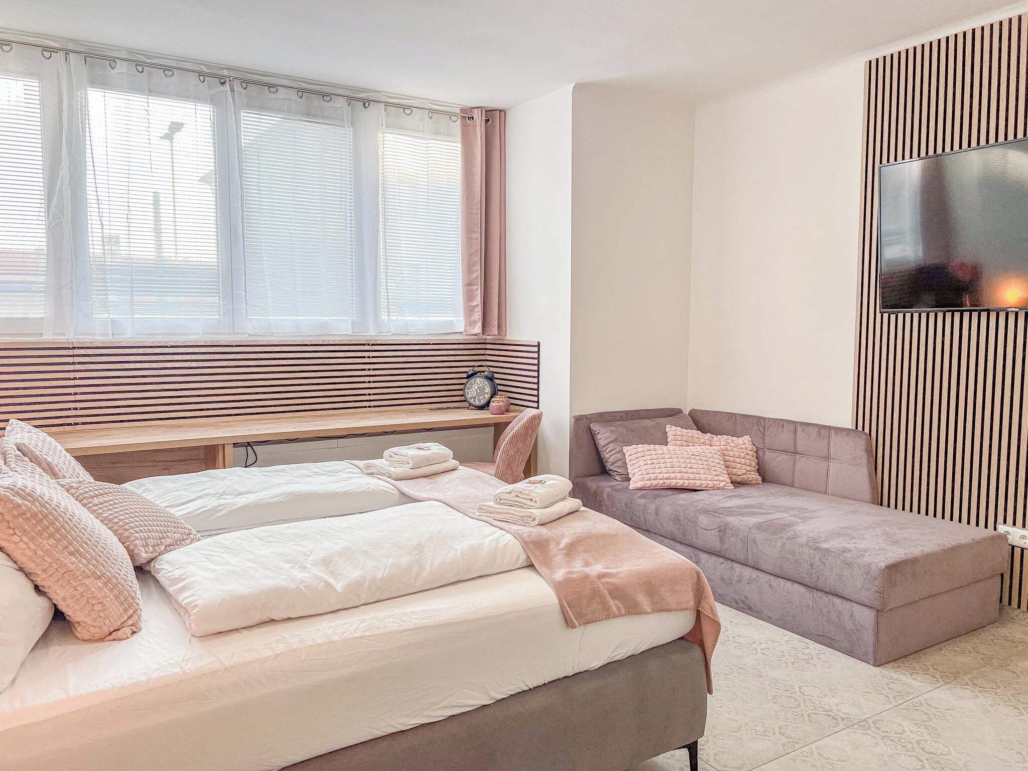 A bedroom with a bed, a bedside table, a nightstand, a window with blinds, and a wall-mounted TV.