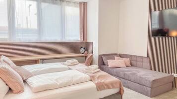 A bedroom with a bed, a bedside table, a nightstand, a window with blinds, and a wall-mounted TV.