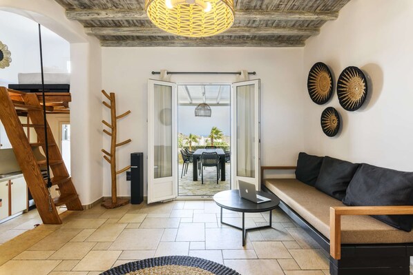 A modern living room with a wooden staircase, a sofa, a dining area with a table and chairs, and a view through a glass door to an outdoor patio. - Aurora Villa 1 (Kalafatis)