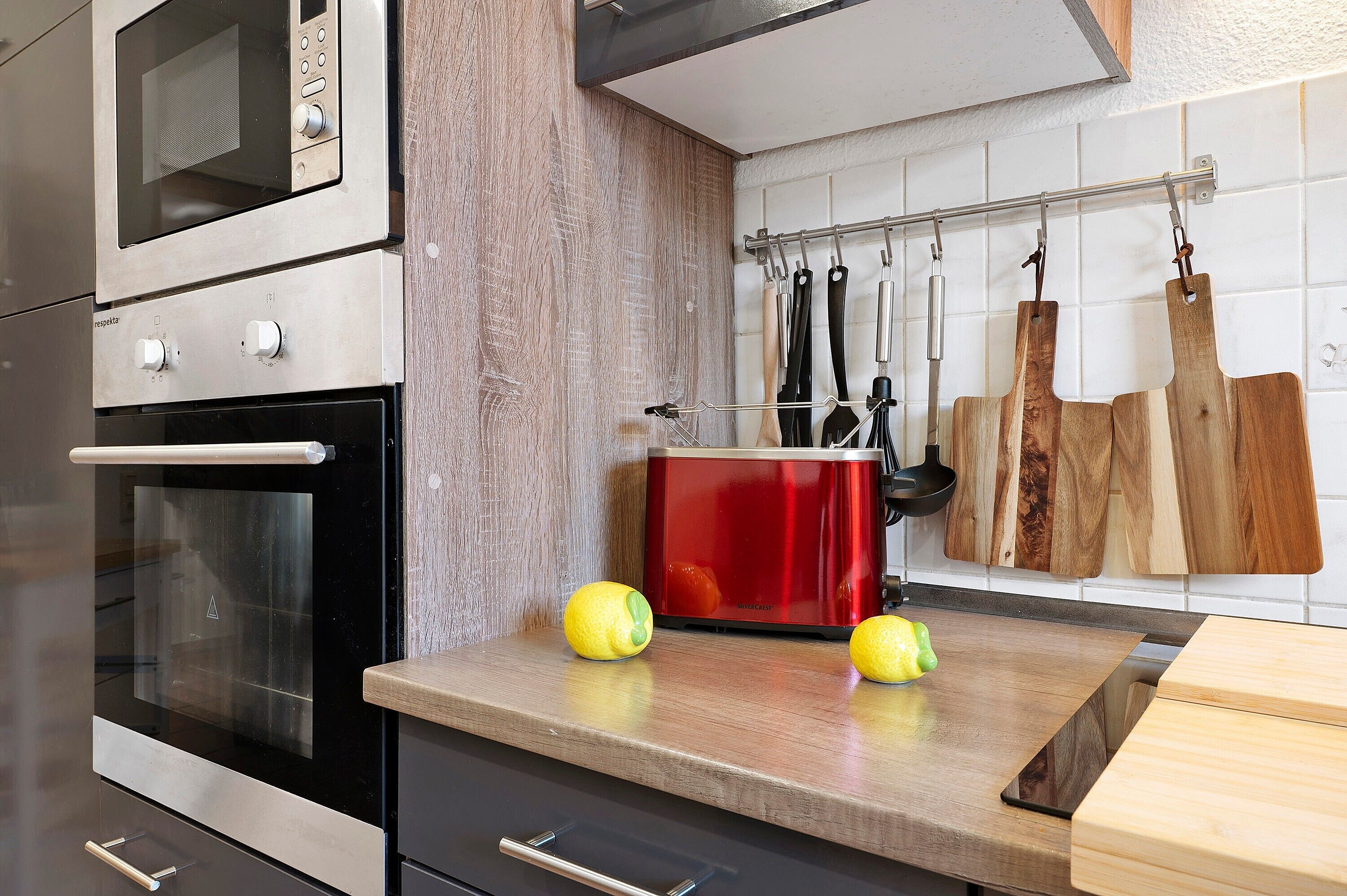 Une cuisine équipée d’un four à micro-ondes, d’un four traditionnel et d’une mijoteuse rouge. On peut voir des citrons sur le plan de travail et des planches à découper en bois accrochées au mur.