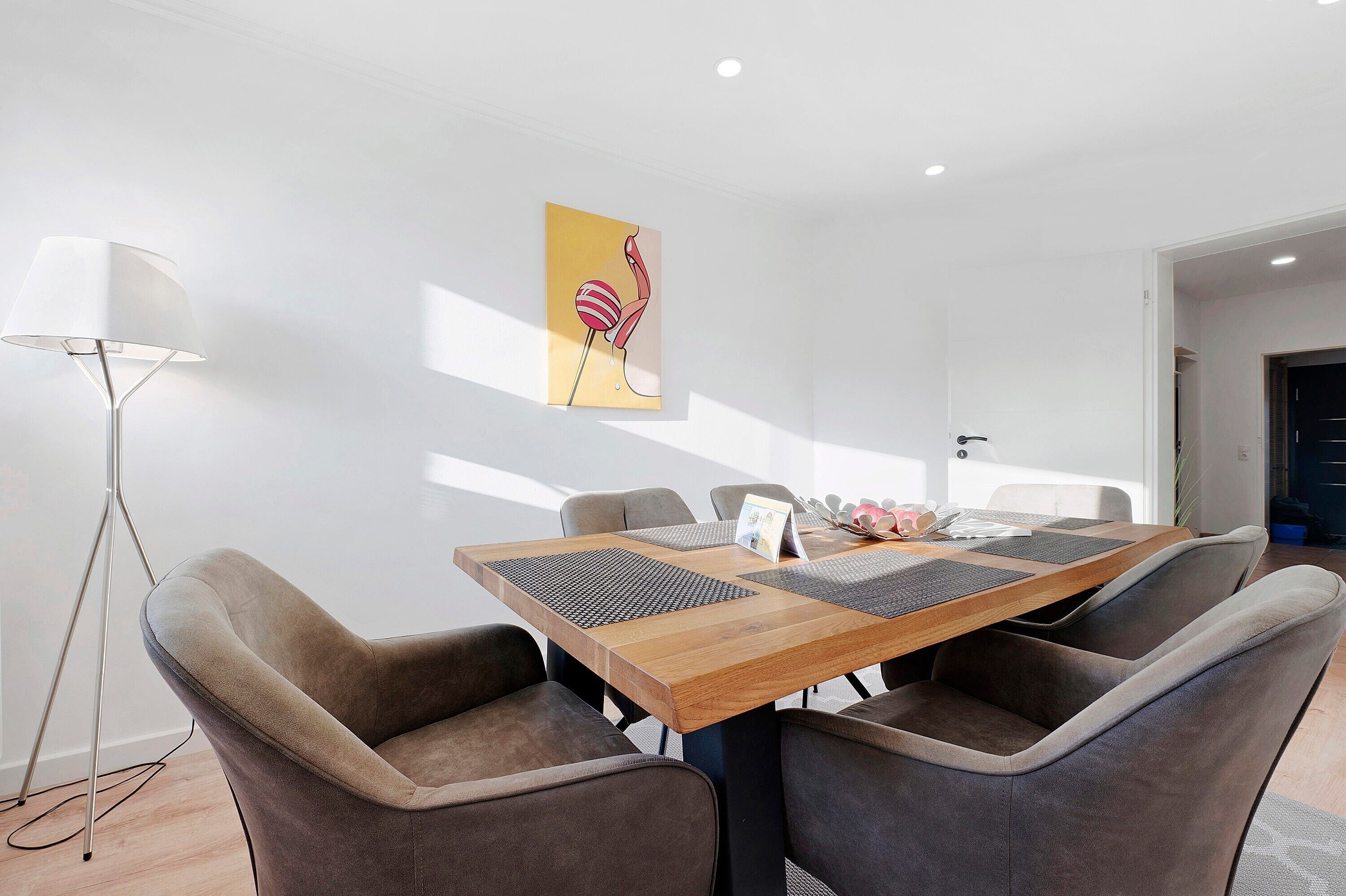 A modern dining area with a wooden table, cushioned chairs, a floor lamp, and a wall-mounted artwork.