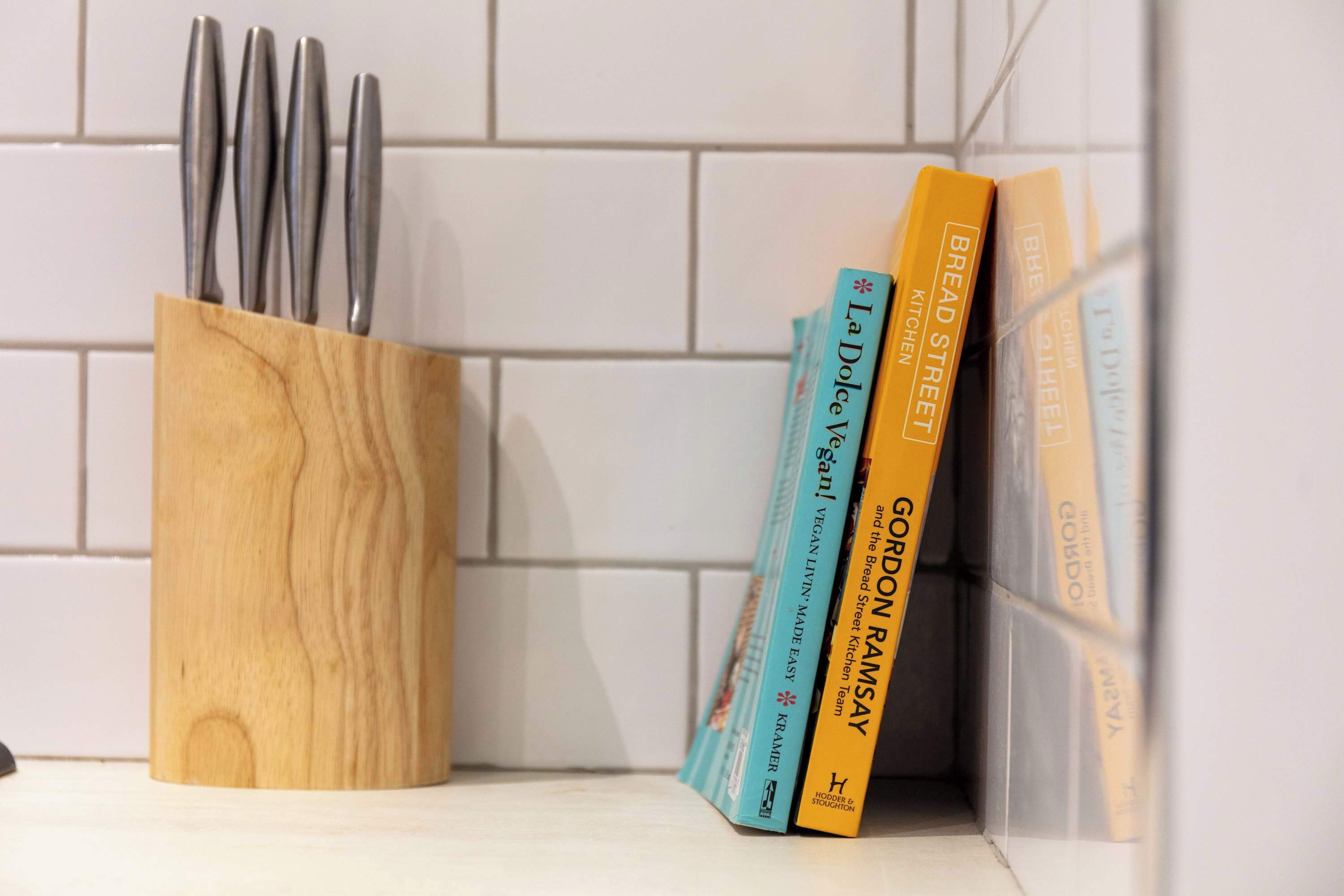 Un soporte de madera para utensilios con utensilios metálicos y tres libros de cocina sobre una superficie de baldosas blancas.