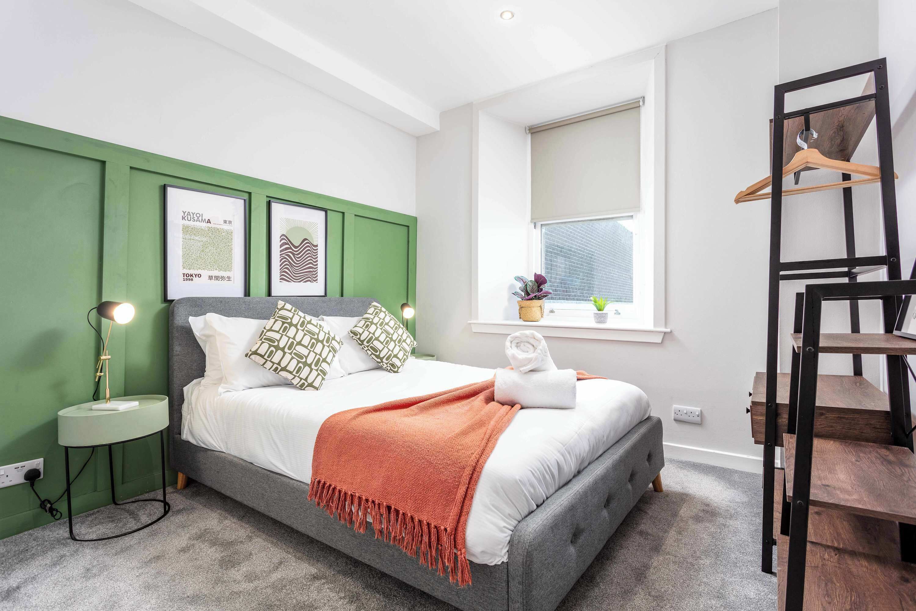 A bedroom with a bed, green walls, a window, a bedside table with a lamp, and a wooden ladder.