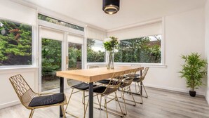 Un comedor con mesa y sillas de madera, ventanas amplias y una planta en maceta.