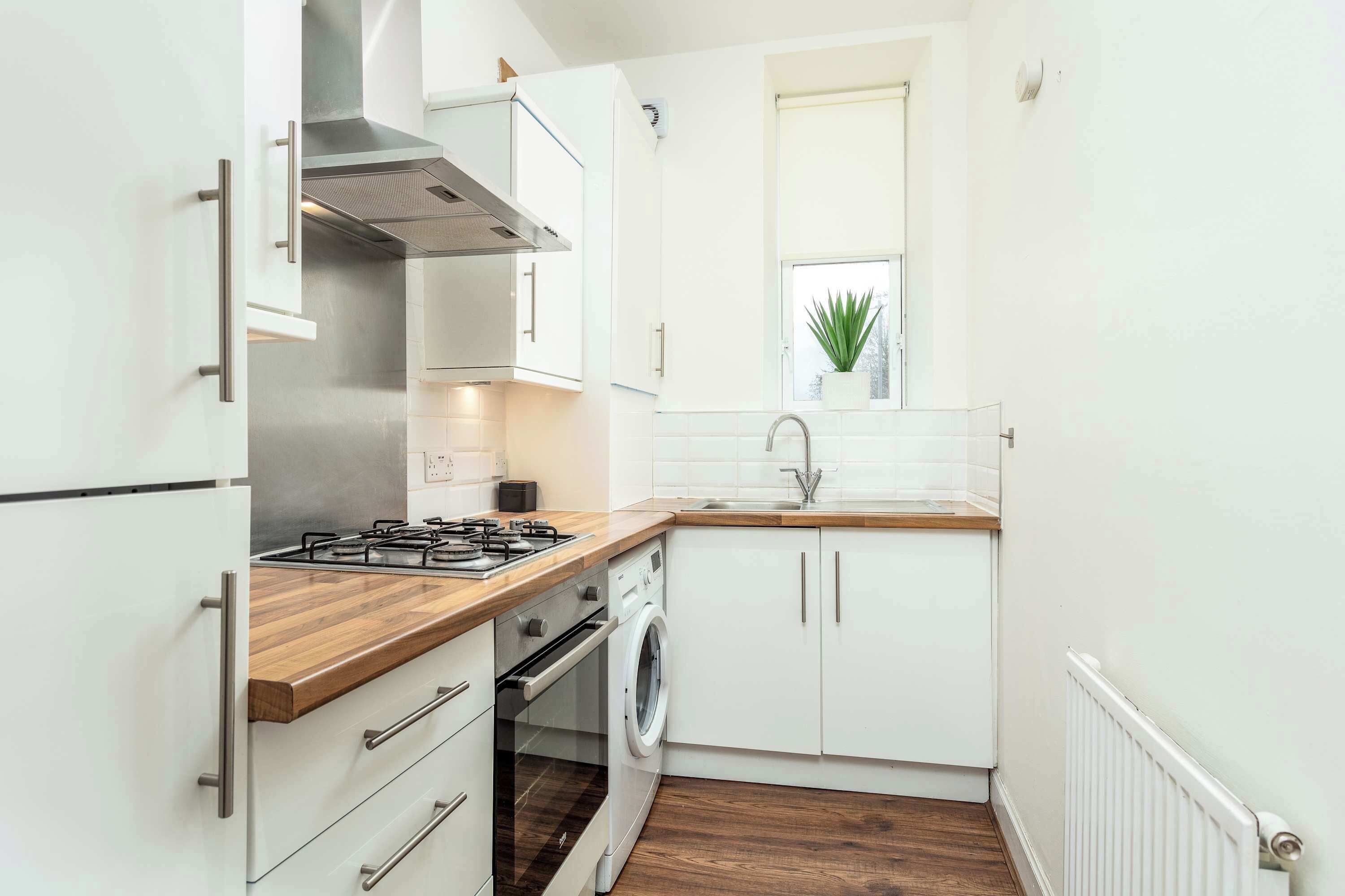 A compact kitchen with white cabinets, a wooden countertop, a built-in oven, a washing machine, and a window with a plant.