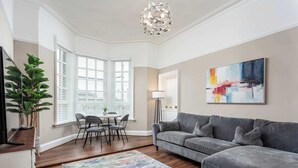 Uma sala de estar com um sofá cinzento, um tapete colorido, uma mesa de jantar com cadeiras, uma planta em vaso e um quadro abstrato grande na parede.