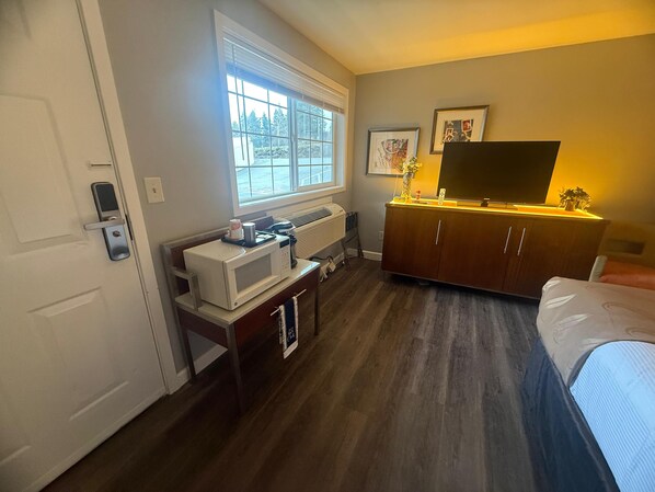 A hotel room with a bed, a desk with a microwave, a dresser, a TV, and a window with blinds. - Naval Suites By FairBridge (Bremerton)