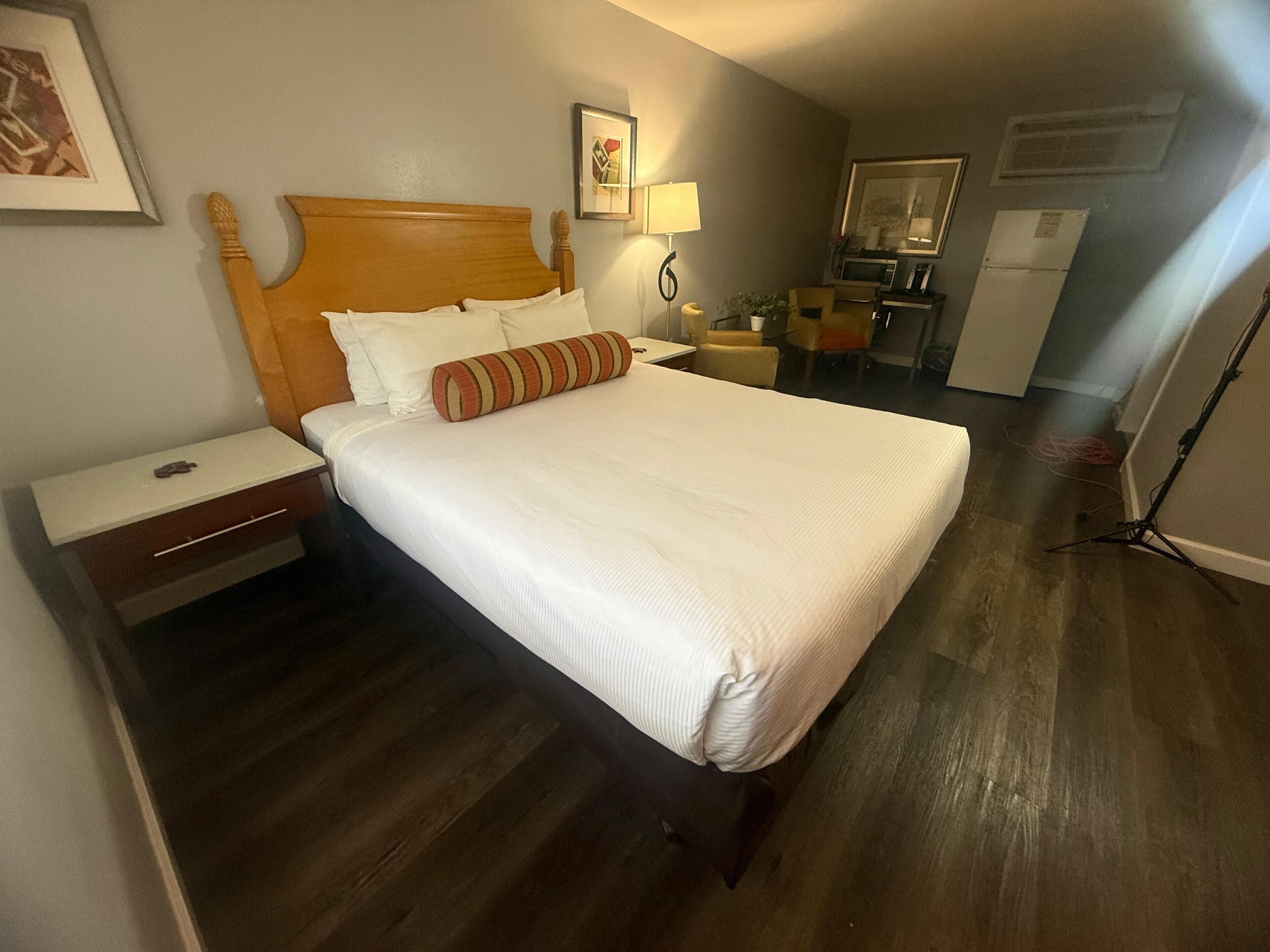 Quarto de hotel com uma cama grande, mesa de cabeceira, abajur, cadeira, escrivaninha, frigorífico e um quadro na parede.
