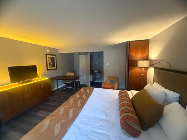 A hotel room with a bed, a desk, a chair, a TV, and a lamp. - Naval Suites By FairBridge (Bremerton)