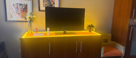 A hotel room with a wooden cabinet, a flat-screen TV, a framed picture, a vase with flowers, and a sign with WiFi details.