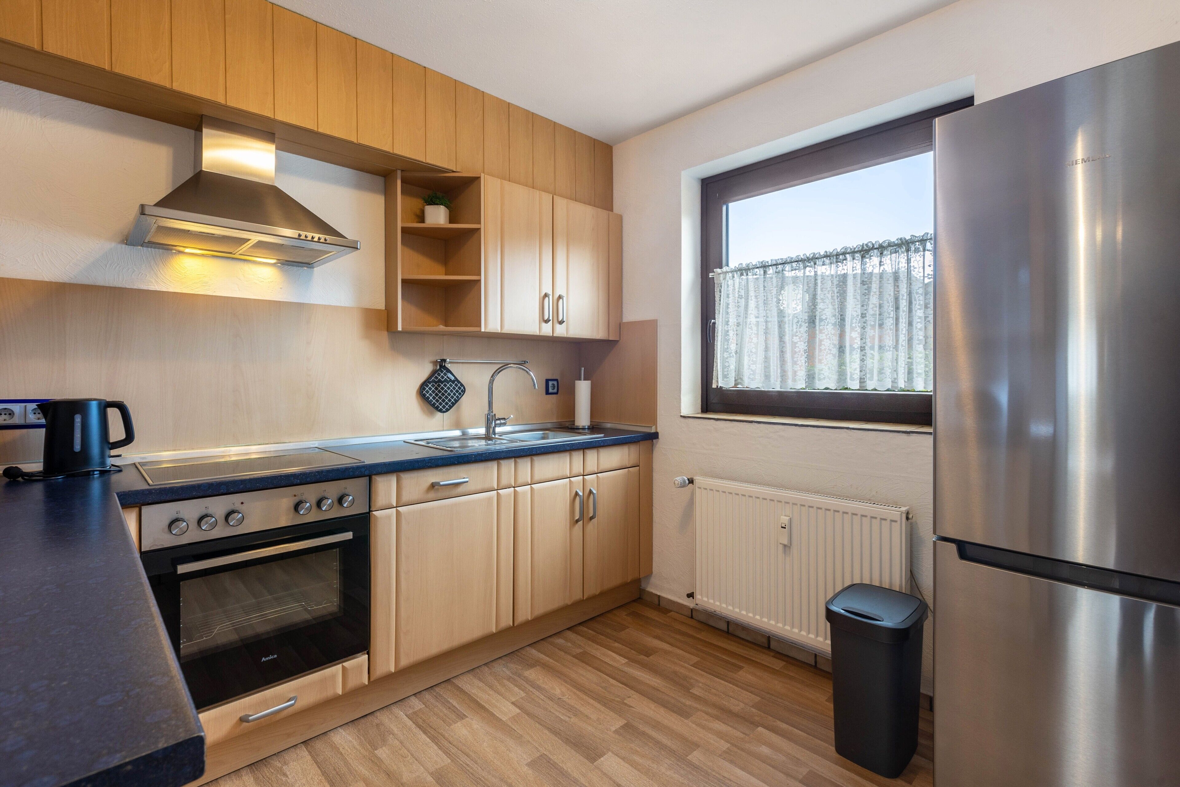 A modern kitchen with wooden cabinets, a stainless steel range hood, a built-in oven, a sink, and a window with a view.
