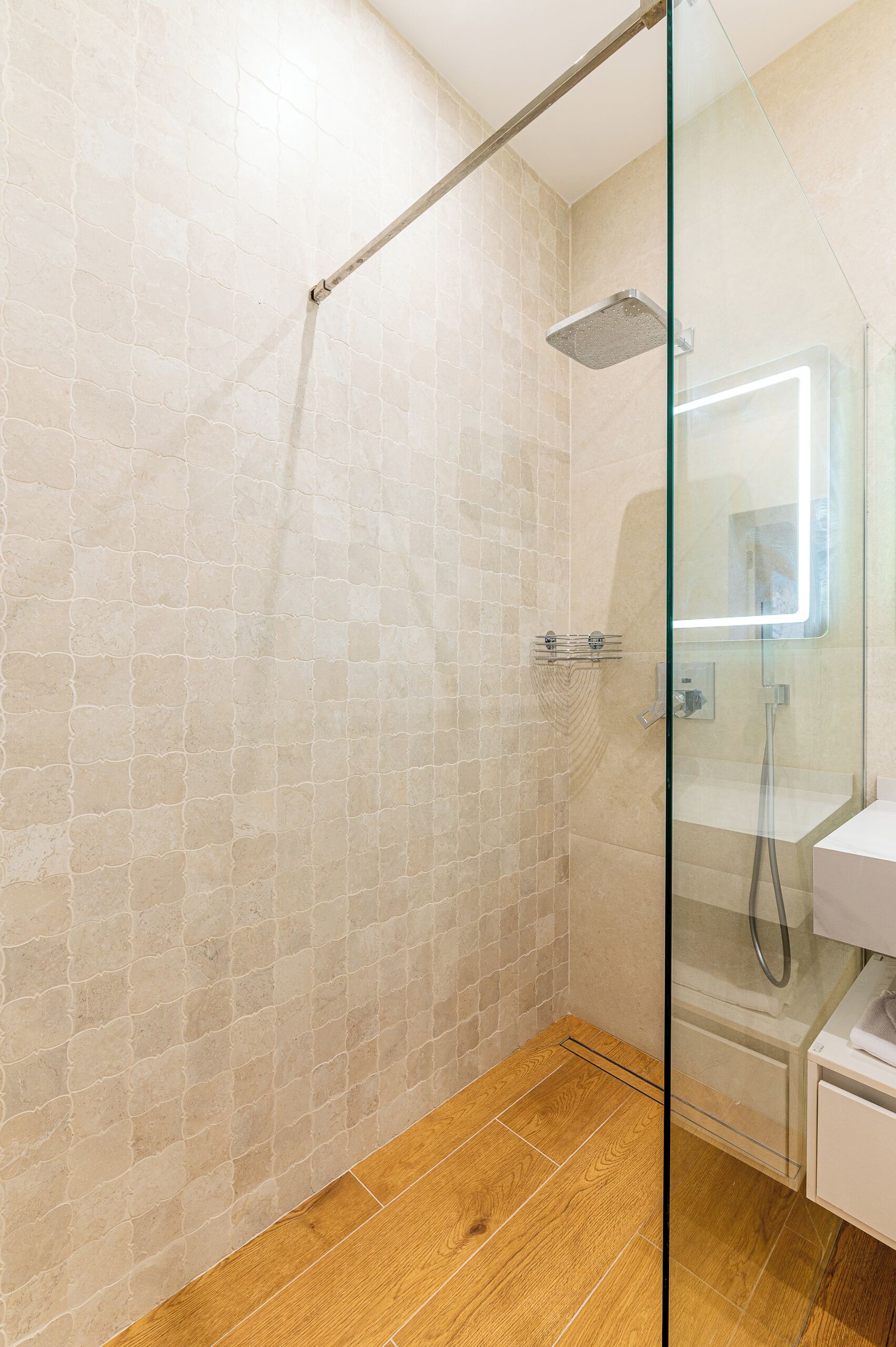 A glass-enclosed shower with a wooden floor, a tiled wall, and a white sink with a mirror.
