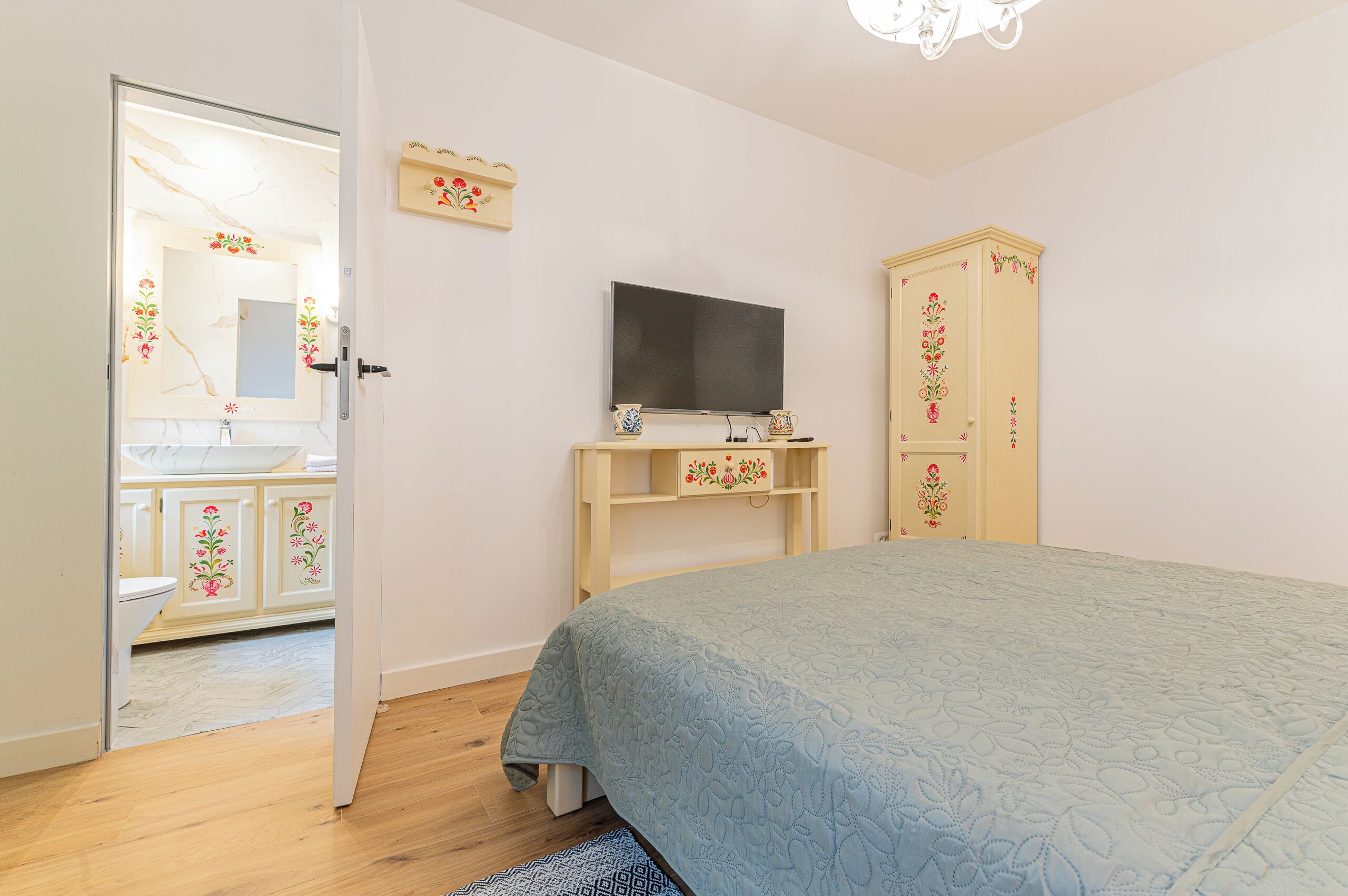 A bedroom with a bed, a television on a stand, a wardrobe, and a bathroom visible through an open door.