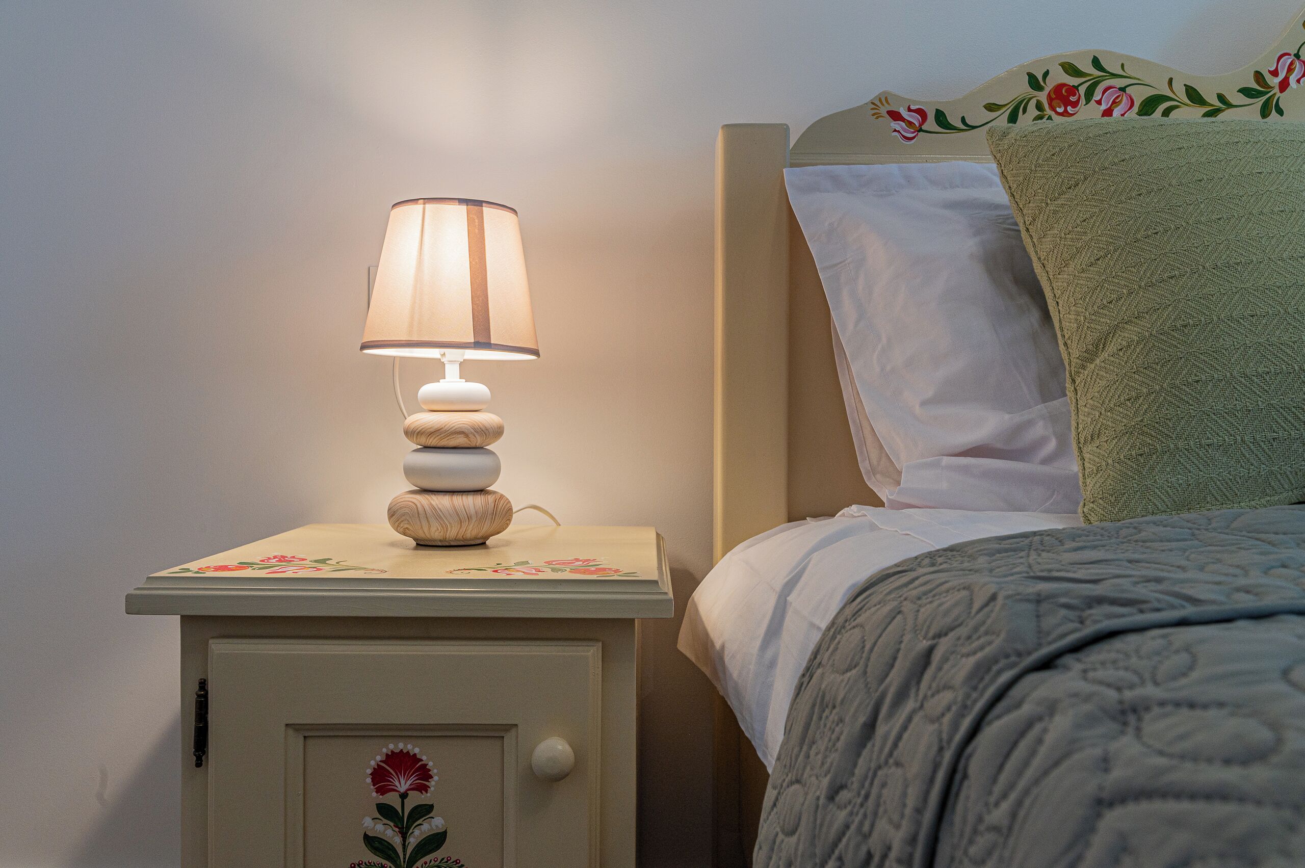 Une table de chevet avec une lampe, un lit recouvert d’une couverture verte et un oreiller à motifs floraux.
