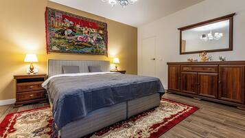 Une chambre avec un grand lit, deux tables de chevet avec des lampes, une armoire en bois, une grande tapisserie au mur et un lustre.