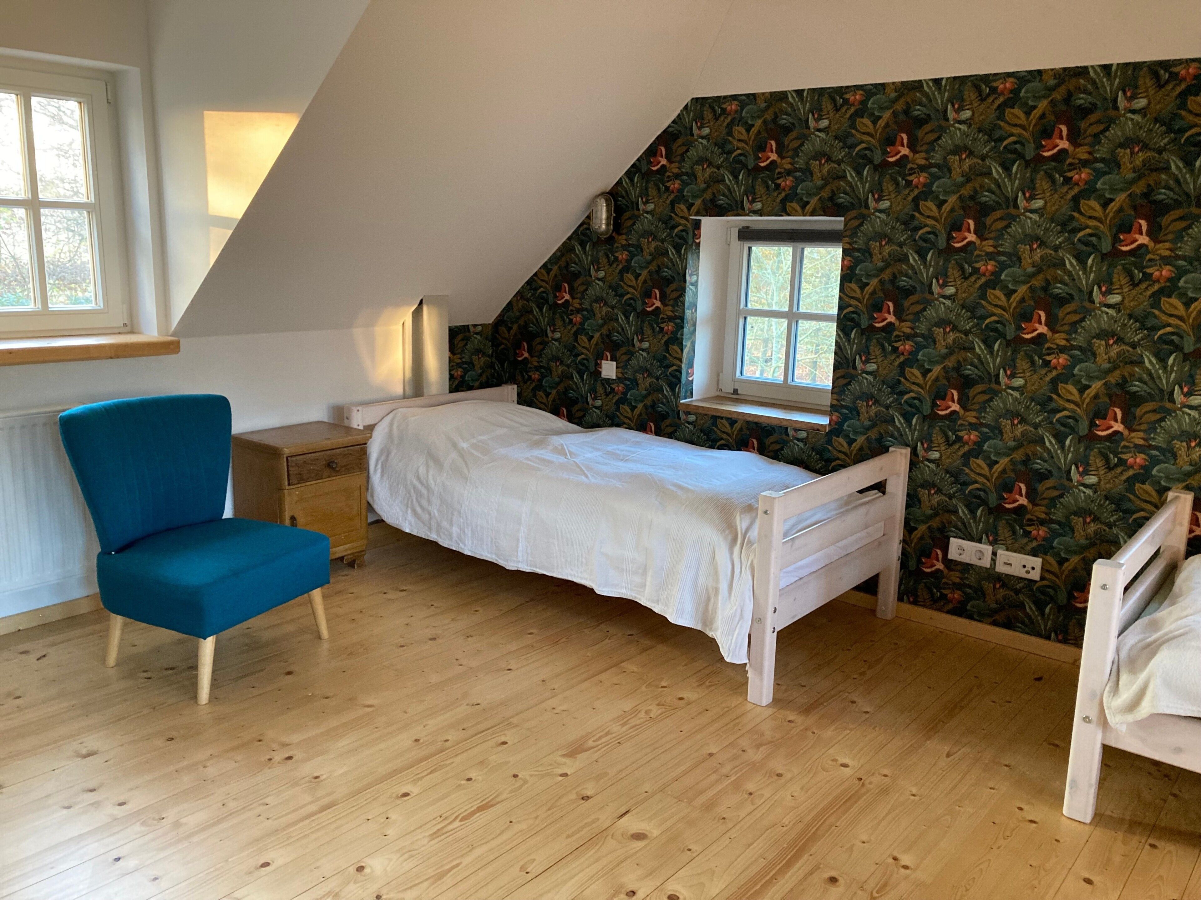 Une chambre avec un plafond en pente, un lit simple, une chaise bleue, une table de chevet, une fenêtre et un mur orné d’un motif de feuilles.