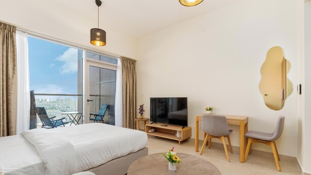 A modern hotel room with a large bed, a TV, a desk with a chair, and a balcony with a view of the city. - Modern Studio in Azizi Samia by Bespoke (Dubai)