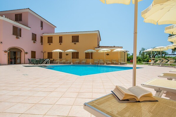 A pool area with lounge chairs and umbrellas, a pool, and a building with multiple windows and balconies. - Comfy Studio With Shared Pool (Orosei)